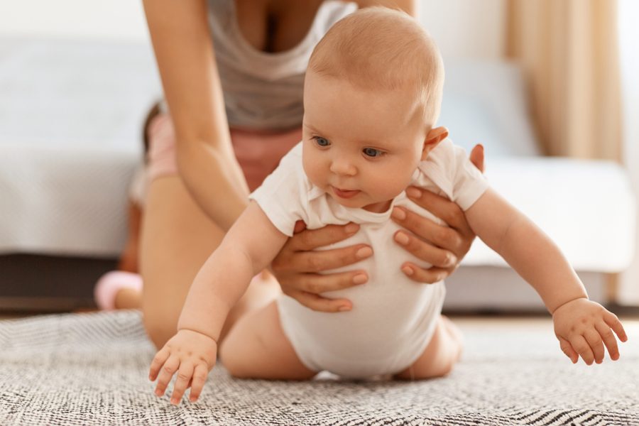 Adorable cute baby in white bodysuit crawling on floor on carpet while mother is helping and supporting, posing in light room at home, happy childhood.