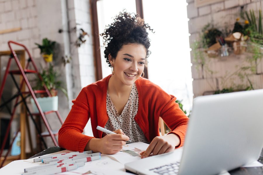 Young smilng woman with dark curly hair sitting at the table happily working on laptop drawing fashion illustrations spending time in modern cozy workshop with big windows