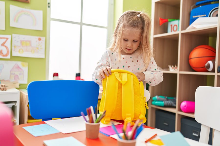 Adorable blonde girl preschool student smiling confident opening backpack at kindergarten