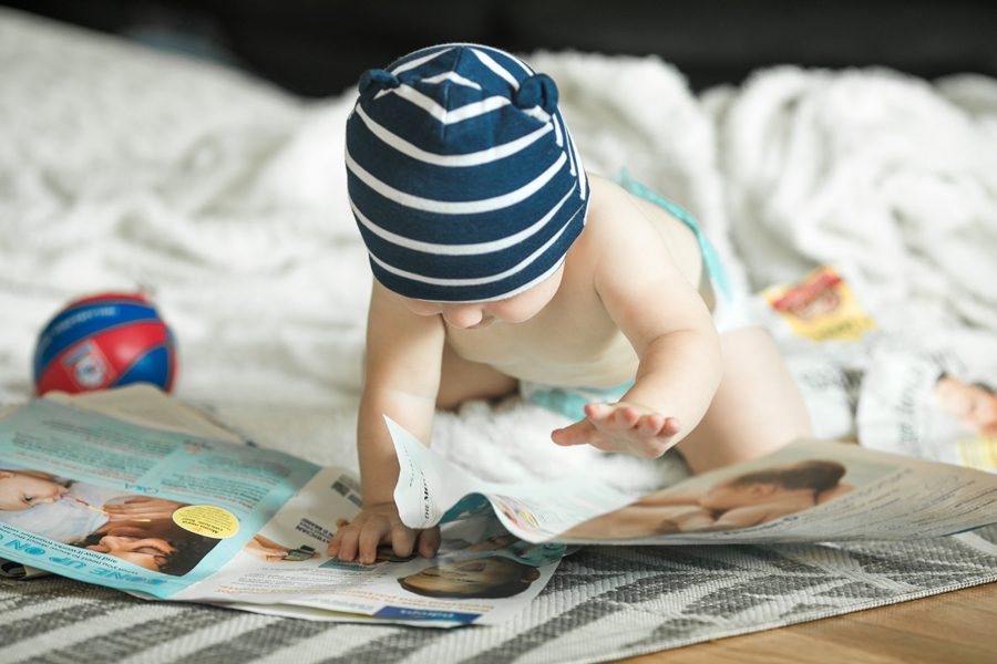 Adorable baby boy learning to crawl and playing with paper magazine in white sunny bedroom. Cute laughing child seating  on a rug. Nursery, clothing and toys for little kids.