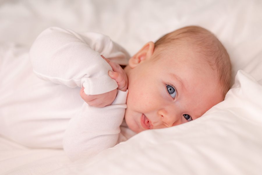 baby with blue eyes in a white bodysuit lies on the back pressing his arms to his chest on white bed linen. happy child. lifestyle. High quality photo