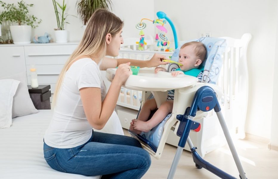 Beautiful young mother feeding her baby boy in highchair