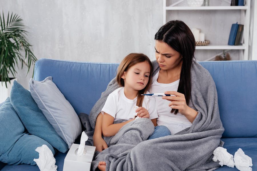 Caring mother holding thermometer in hand and frowning while hugging her daughter