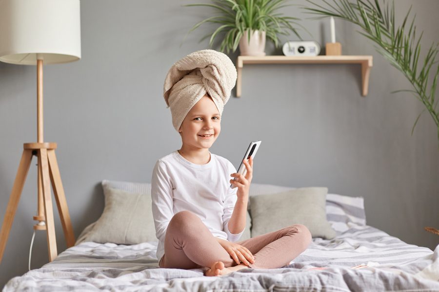 Cheerful joyful little female kid in towel sitting in bed in bedroom after taking shower using smart phone weekend morning looking at camera.