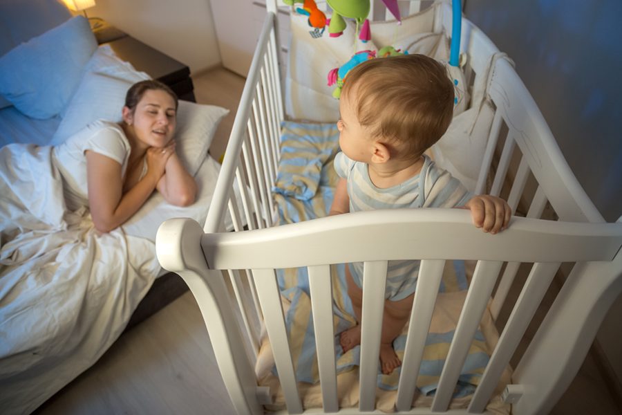 Cute baby standing in crib and looking at tired mother that fell asleep