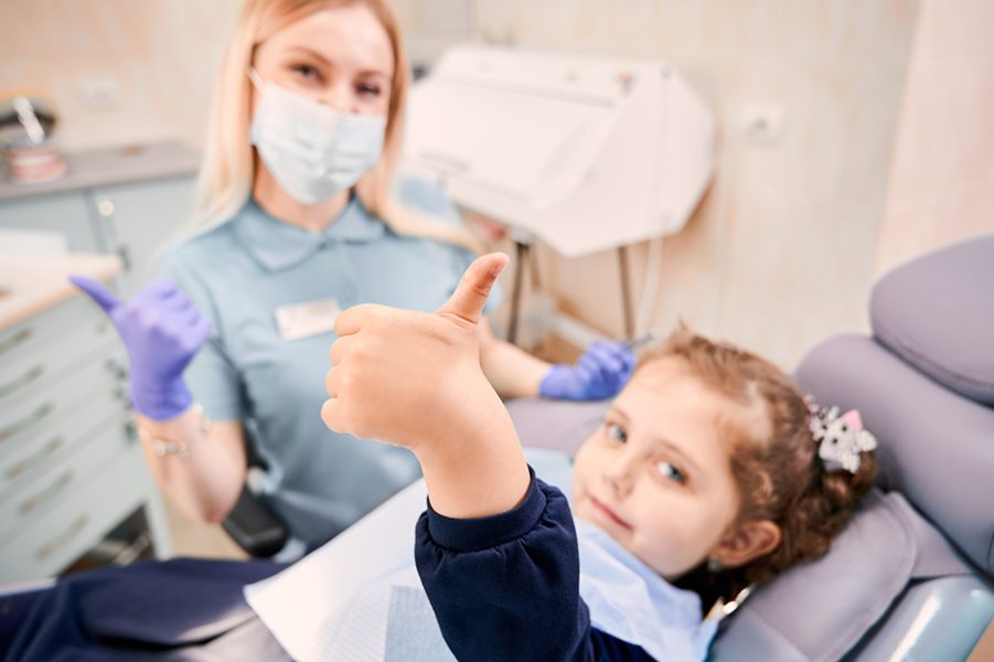Focus on girl's hand giving thumbs up while woman dentist in medical mask sitting beside kid, showing approval gesture after dental procedure. Concept of pediatric dentistry and dental care approval.