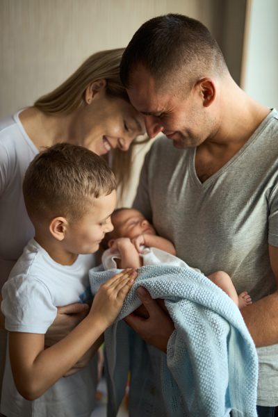 Father and mother holding newborn daughter in arms, older brother stands next to and looks with love. Family values