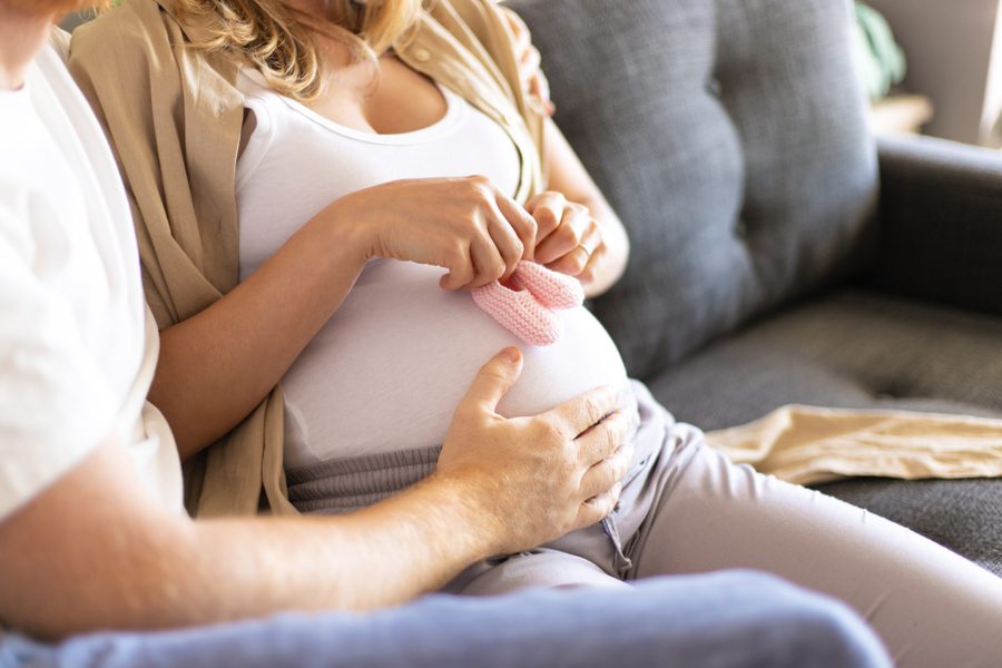 Future mother applying baby girl shoes to belly. Her husband sitting on couch, embracing wife and touching belly. Pregnant couple at home concept
