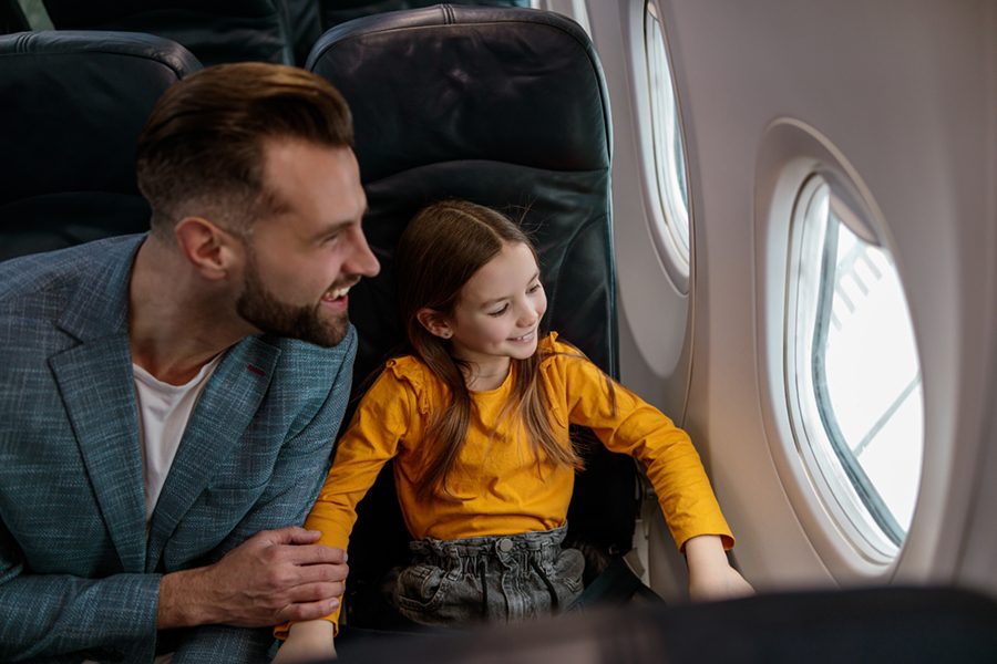 Cheerful little girl enjoying view from aircraft window and smiling while sitting next to dad in passenger chair