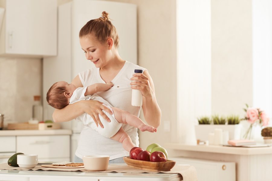 Happy young mother in white Tshirt feeding her baby with baby formula standing in kitchen