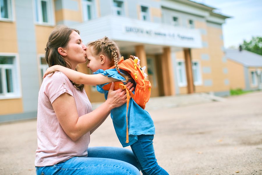woman morally supports her daughter by holding her hands and kissing her forehead. mother accompanies student to school. friendly family goes to kindergarten.happy little girl with a caring mother.