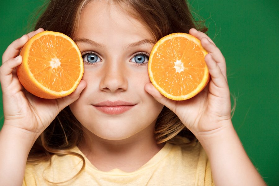Young pretty girl holding orange, looking at camera, smiling over green background.