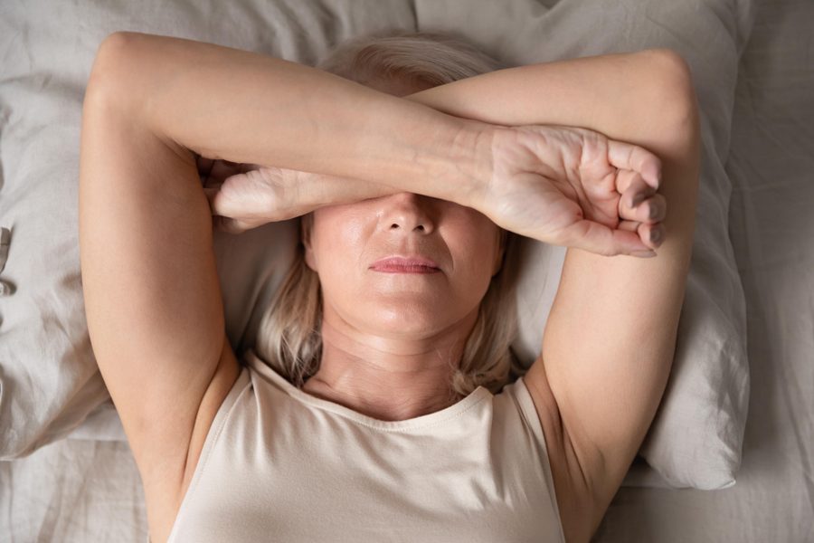 Sleepless mature woman suffering from insomnia close up, lying in bed, older female covering eyes with hands, trying to sleep, nightmares or depression, feeling headache or migraine