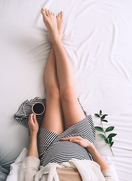 Beautiful pregnant woman holding her belly and relaxing with cup of morning coffee in bed during pregnancy. Young mother waiting for baby. Minimalist modern style photography.