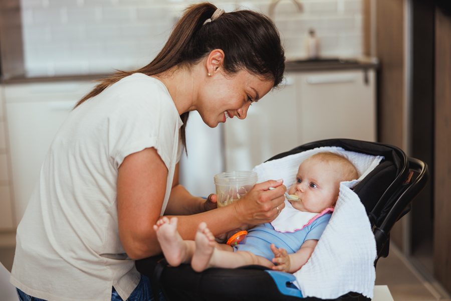 Shot a smiling woman sitting in the kitchen with her adorable baby and feeding her.