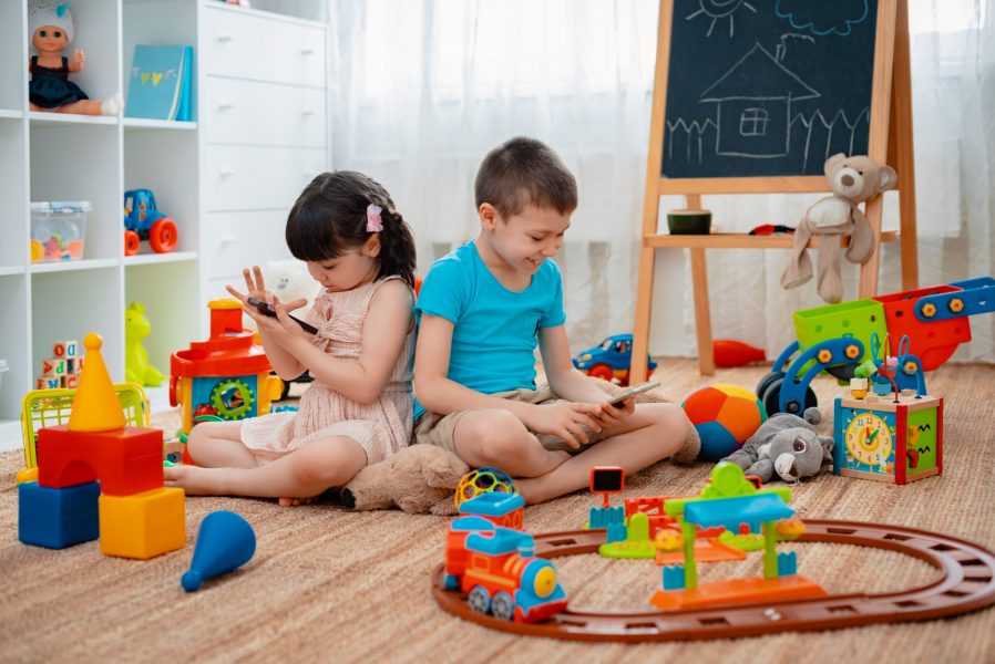 Siblings children brother and sister, friends sit on the floor of the house in the children's play room with smartphones, detached from the scattered toys. Concept of new gadgets for kids.