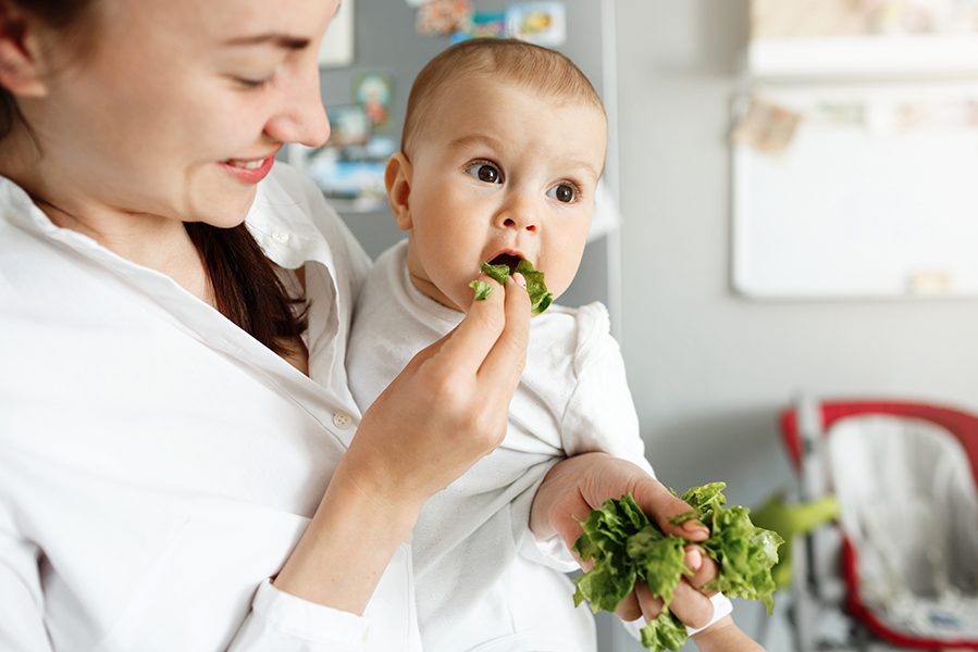 Close up of mother feeding her little baby with salad leaves during cooking lunch for whole family. Child looking aside and happily eats lettuce