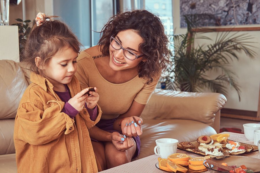 Smiling young mother with cute little daughter having breakfast on the sofa at home.