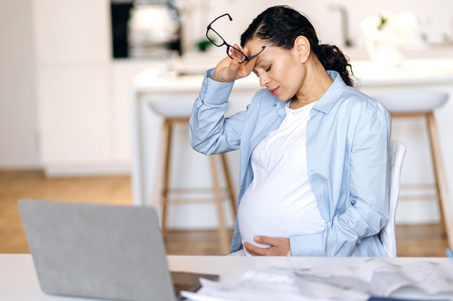 Fatigue, stressful mixed race mature pregnant woman, freelance worker, designer or manager working remotely, sits at desk, taking break, having headache, holding glasses in hand, eyes closed