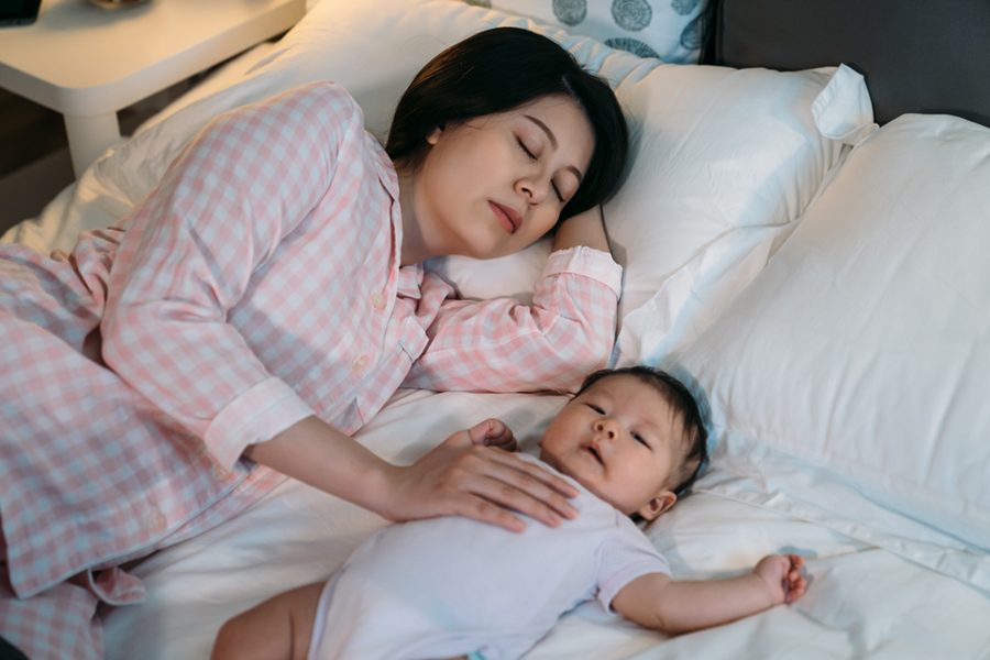 tired asian first time mom keeping hand on the waking baby’s chest is falling asleep while putting it to sleep on bed in the bedroom at night.