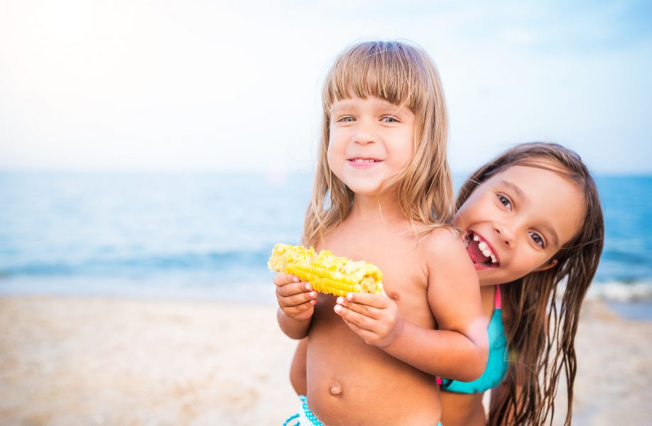 Two kids having fun on beach, smiling and eating corn. Girls looking at camera, portrait on blurry background