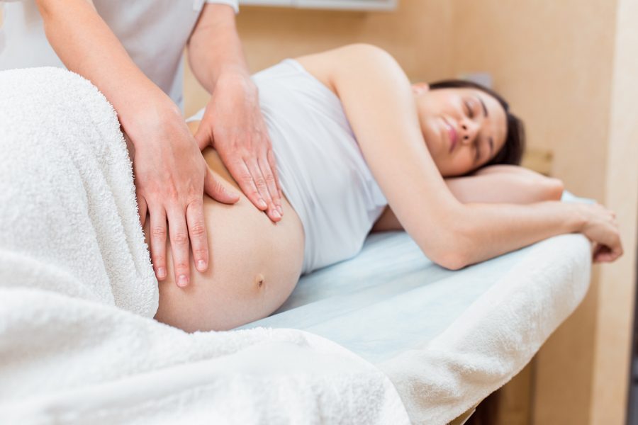 A beautiful pregnant brunette with long hair lies on her side at a reception with a massage therapist in a cosmetology room