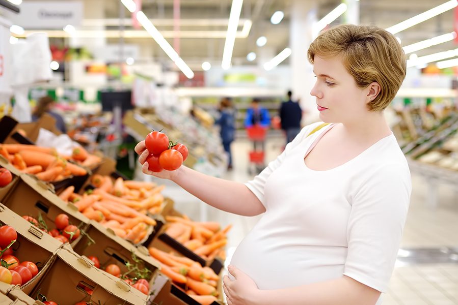 Young pregnant woman choosing fresh tomato in supermarket. Pregnancy and shopping.